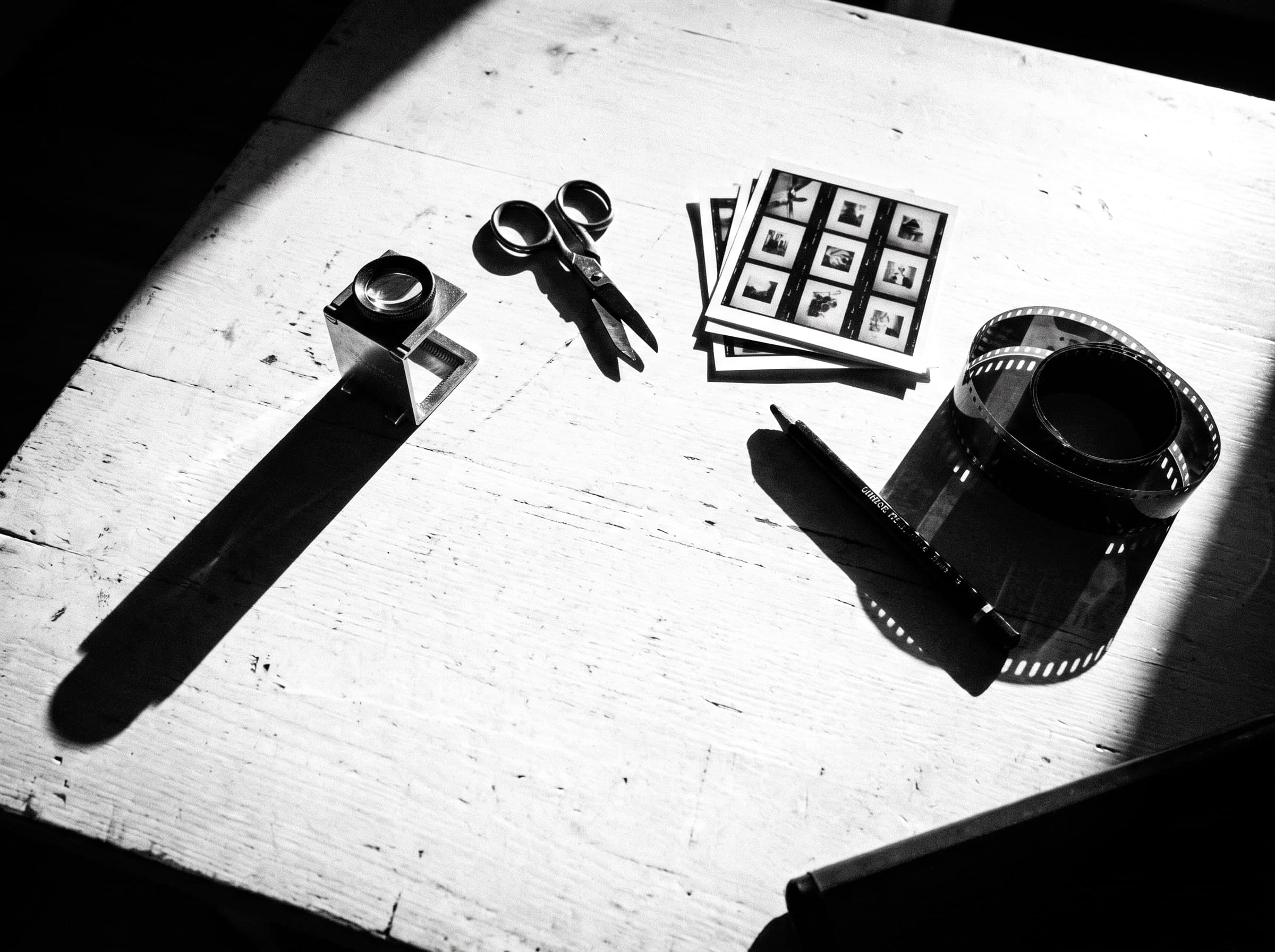 Arbeitstisch von oben: Fadenzähler, Schere, ein Stapel Kontaktbögen, ein Stift und ein aufgerollter Filmstreifen, harte Schatten