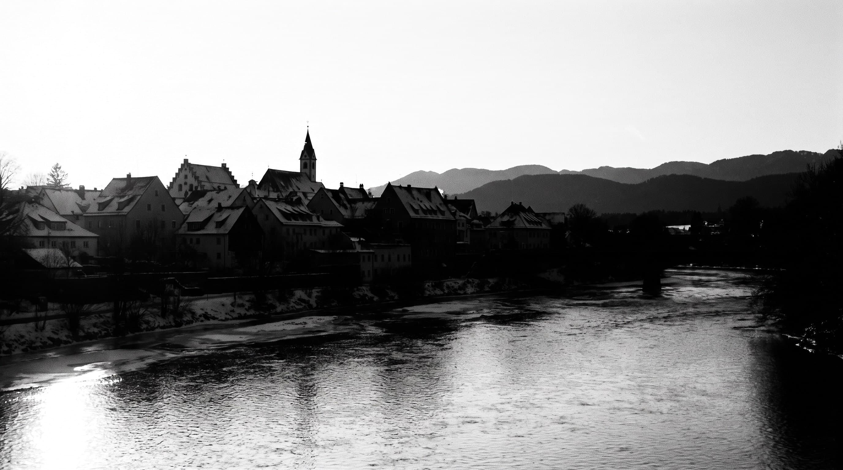 Fluss unterhalb einer Altstadt, Giebelreihen und Kirchturm als schwarze Silhouette, Wasseroberfläche im Gegenlicht ausgebrannt