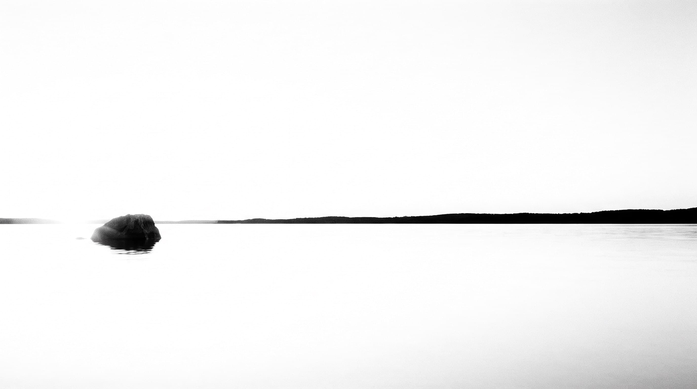 Stilles Wasser bis zum Horizont, links bricht ein einzelner dunkler Fels die Fläche, rechts eine flache dunkle Uferlinie