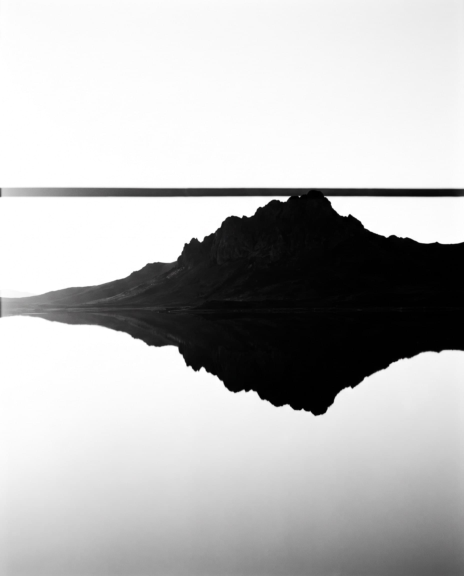 Dunkler Berg, exakt gespiegelt in vollkommen stillem Wasser, heller Himmel