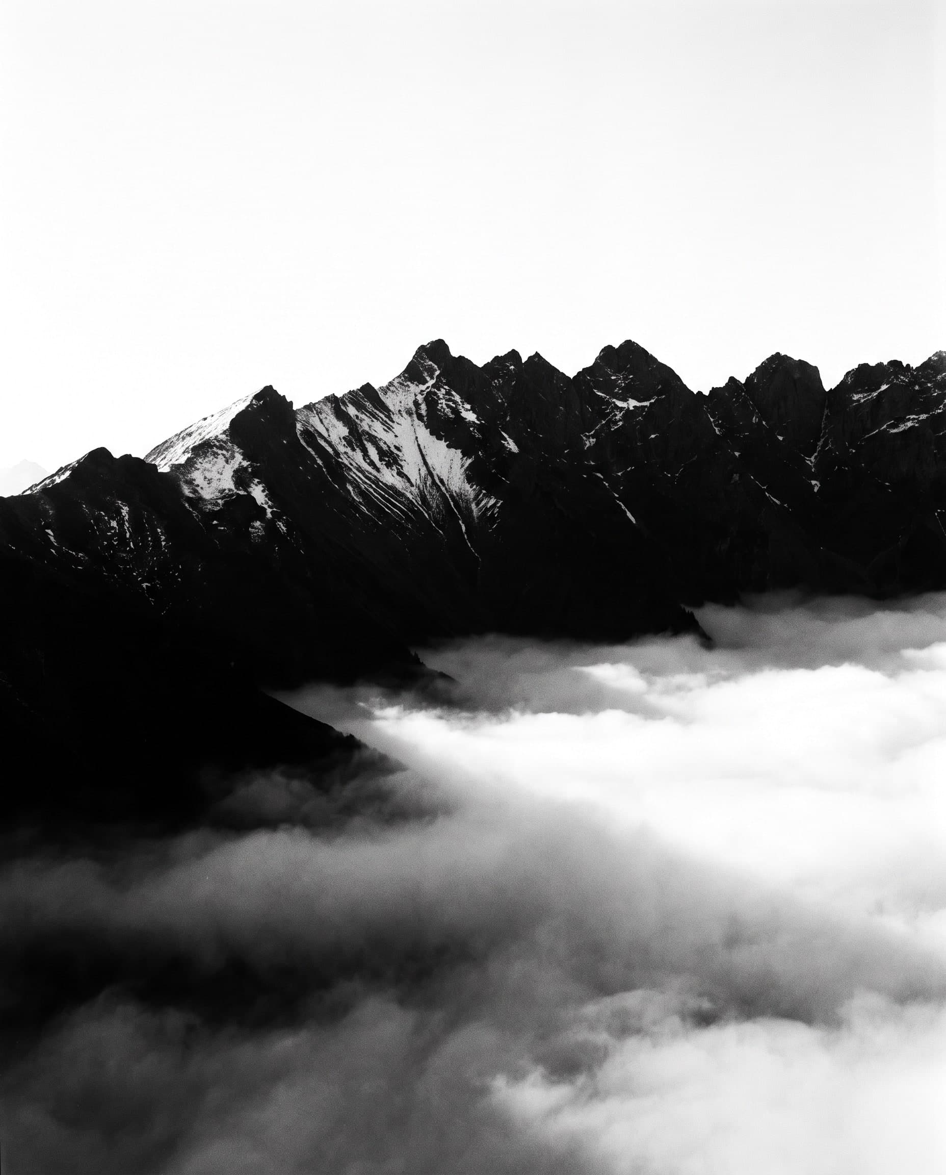 Alpiner Grat als harte schwarze Silhouette über Nebelbänken, Himmel nahezu papierweiss