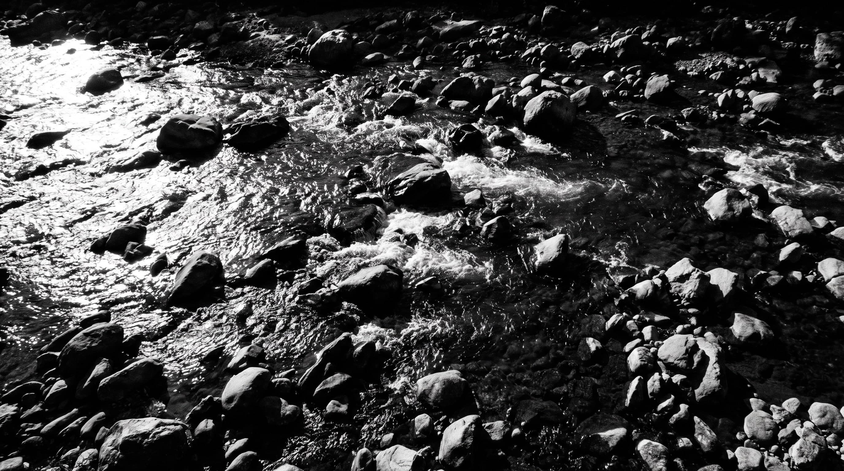 Steiniges Flussbett von oben, dunkle Blöcke, zwischen denen Wasser in hellen Bahnen aufbricht